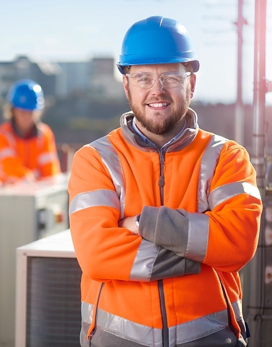 An air conditioning engineer is finishing the installation of several units on a rooftop. Two colleagues can be seen also installing units in the background. They are wearing hi vis jackets, hard hats and safety goggles.